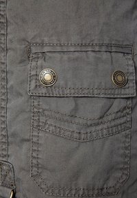 Gros plan sur une poche de veste en tissu gris avec deux boutons métalliques portant l’inscription "Tom Tailor" et des coutures décoratives visibles.
