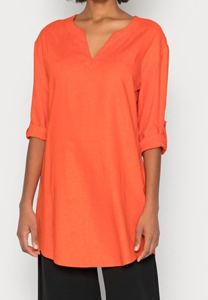 Oranje linnen tuniek met een V-hals, driekwart opgerolde mouwen en een asymmetrische zoom, gecombineerd met donkere broeken.