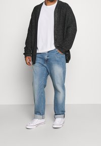 Mörk stickad kofta över en vit t-shirt, kombinerad med ljusblå jeans och vita sneakers. Kläders texturer synliga, avslappnad passform.