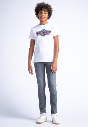 Witte katoenen t-shirt met blauwe en rode "PETROLIND" grafiek, grijze skinny jeans en witte sneakers met lichte accenten. Krullend haar.