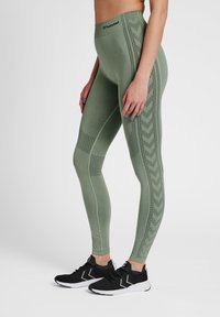 Leggings de sport verts avec une taille haute, des motifs texturés sur les côtés et une coupe ajustée, portés avec des baskets noires.