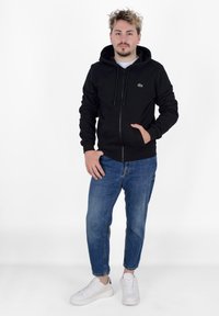 Sudadera con cremallera negra, con un bolsillo canguro, puños elásticos y un pequeño logo verde. Combinada con unos jeans ajustados de color azul claro y zapatillas blancas.