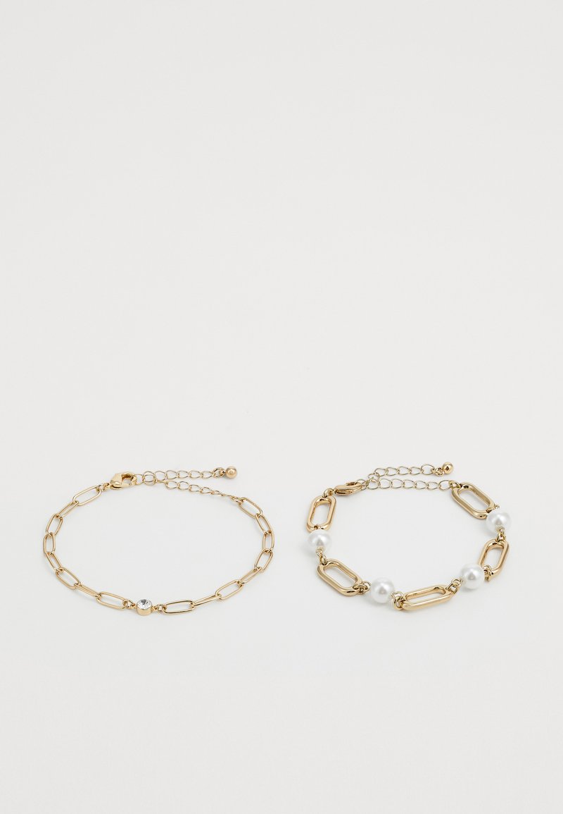 Duas pulseiras de corrente em ouro sobre fundo branco; uma com uma pequena gema transparente, a outra com elos ovais e pérolas brancas.