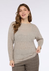 Beżowy dziergany sweter o szerokim dekolcie, z poziomymi prążkowanymi wzorami i subtelnymi zdobieniami z koralików. Zestawiony z kraciastymi spodniami.