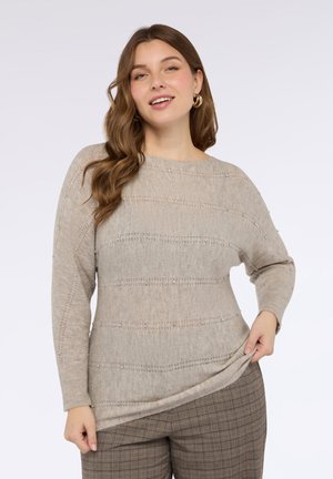 Beżowy dziergany sweter o szerokim dekolcie, z poziomymi prążkowanymi wzorami i subtelnymi zdobieniami z koralików. Zestawiony z kraciastymi spodniami.