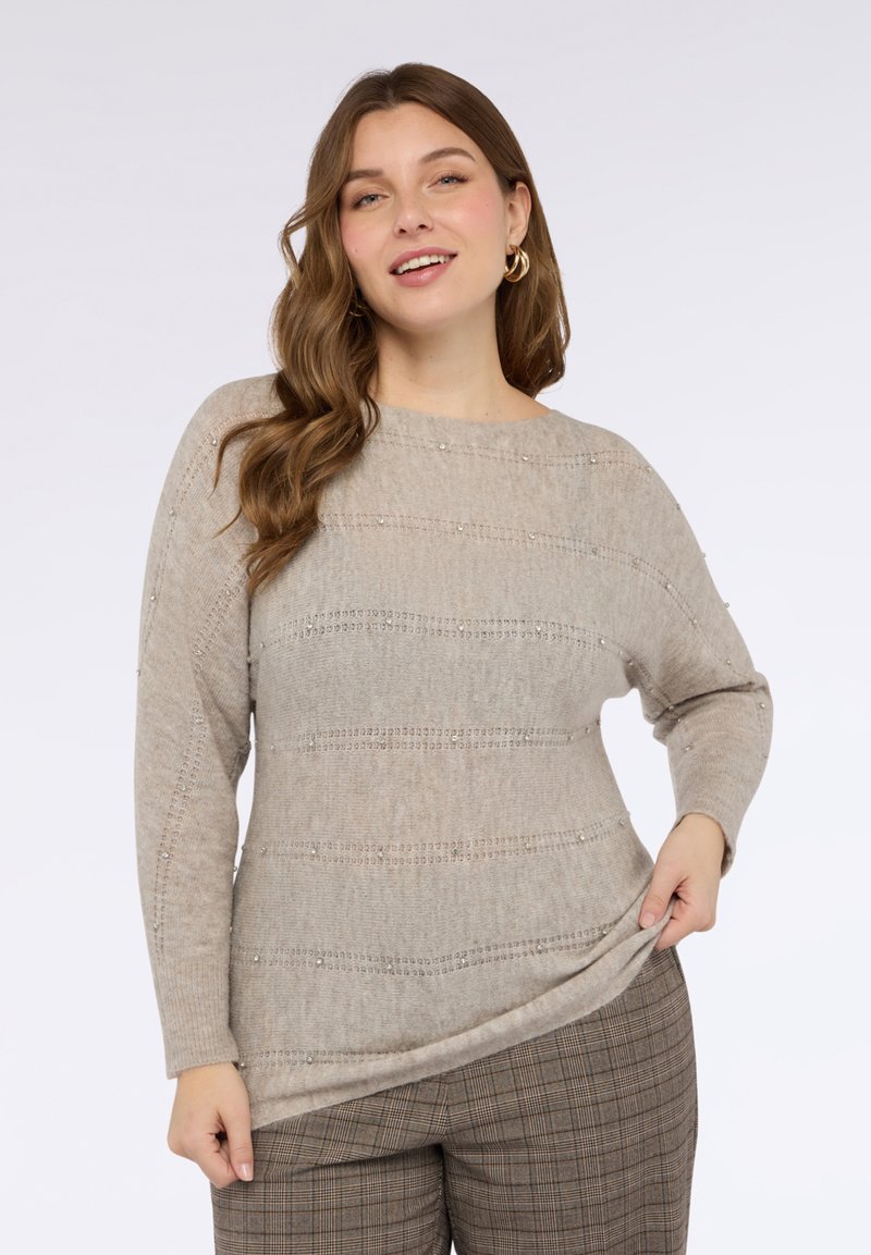 Beżowy dziergany sweter o szerokim dekolcie, z poziomymi prążkowanymi wzorami i subtelnymi zdobieniami z koralików. Zestawiony z kraciastymi spodniami.