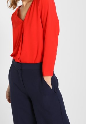 Mujer que lleva una blusa de manga larga de color rojo brillante metida dentro de unos pantalones de talle alto de color azul marino oscuro, con una mano en el bolsillo.