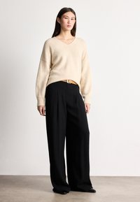 Béžový pletený sveter s véčkovým výstrihom, voľného strihu a rebrovými manžetami, doplnený o čierne široké nohavice s vysokým pásom a hnedý opasok.