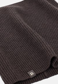 Brūns adīts šalle ar ribotu tekstūru, kurai ir neliela zelta akcentēta logotipa birka. Mīksts materiāls ar gludu apdari, salocīta uz izstādi.