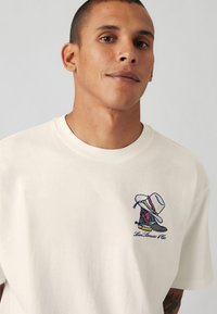 Cream-colored cotton t-shirt featuring an embroidered graphic of a cowboy boot and hat, with colorful accents and logo.