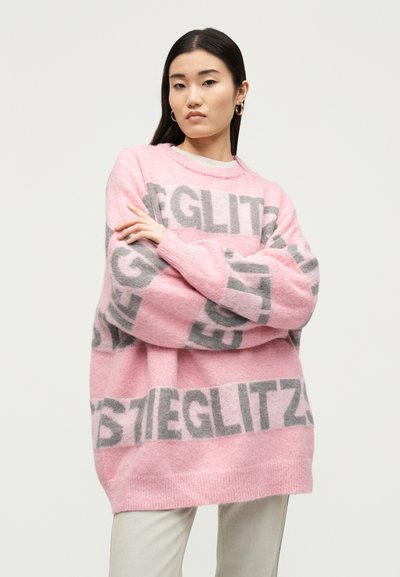 Jeune femme aux longs cheveux foncés porte un pull rose oversize avec de grandes lettres grises et un pantalon blanc, debout les bras croisés.