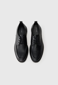 Zapatos de cuero negros con un acabado liso, punta redondeada y diseño de cordones tradicional. Presentan una suela ligeramente elevada y una lengüeta para facilitar el calzado.