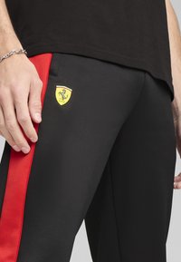 Schwarze Jogginghosen mit roten Seitenstreifen, ausgestattet mit einem gelben Ferrari-Logo. Glatte Textur, taillierte Passform und elastischer Bund sichtbar.