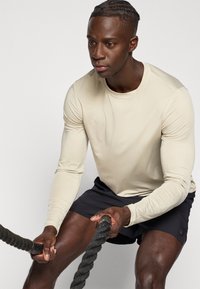 Μπεζ αθλητική μπλούζα μακρυμάνικη με στρογγυλή λαιμόκοψη, σε συνδυασμό με σκούρο σορτς. Το άτομο κρατάει ένα χοντρό, υφασμάτινο σχοινί.