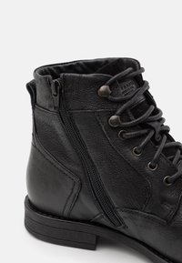 Bottine en cuir noir avec surface texturée, laçage sur le devant, fermeture éclair latérale et talon bas empilé. Marque discrète sur la languette.