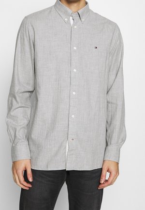 Mężczyzna ubrany w jasnoszarą, zapinaną na guziki koszulę z długim rękawem z małym logo na piersi i ciemne spodnie, stojący na jednolitym tle.