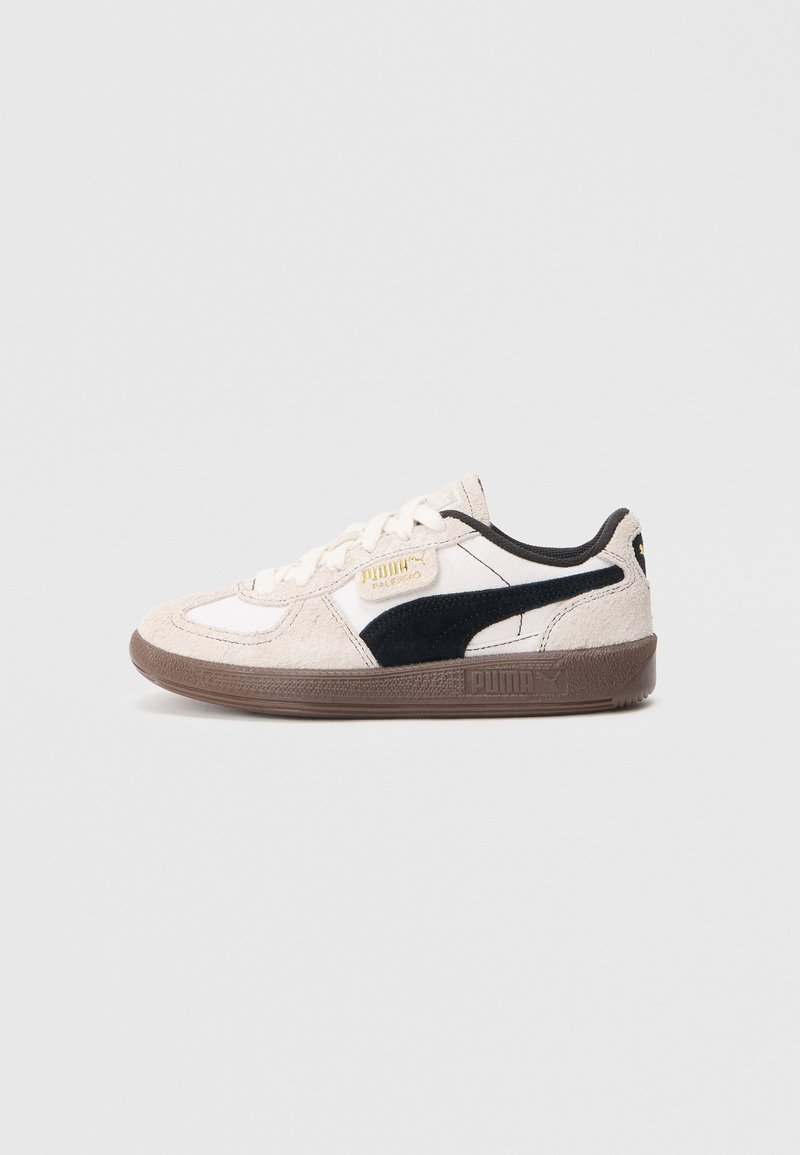Zapatilla con una parte superior sintética en color crema, acentos de gamuza negra y suela de goma marrón. Presenta cordones blancos y detalles en dorado del logo.