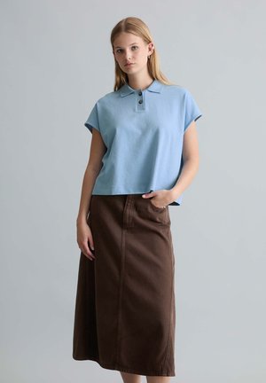Młoda kobieta z długimi blond włosami nosi jasnoniebieską koszulkę polo z krótkim rękawem i długą brązową spódnicę, pozując z jedną ręką w kieszeni.