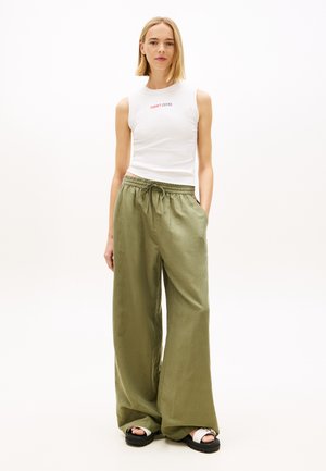 Donna con capelli biondi lisci indossa un top bianco senza maniche Tommy Jeans, pantaloni verde oliva larghi e sandali bianco e nero, in piedi rilassata.