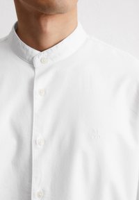 Witte katoenen shirt met een staande kraag, knoopsluiting en een subtiel reliëflogo aan de linkerzijde. Gladde textuur, klassiek ontwerp.