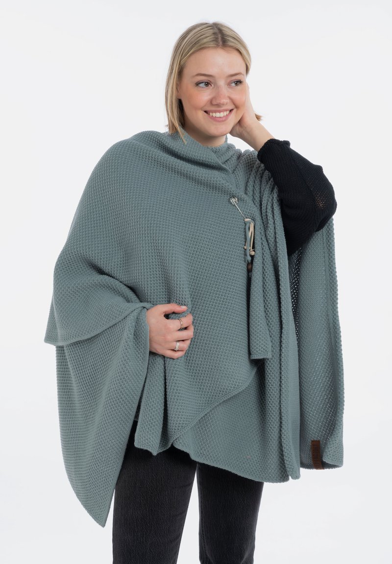 Poncho en tricot gris clair avec un motif texturé, doté d'une fermeture à boutons sur le côté et d'un ourlet asymétrique. Accentué par une étiquette marron.