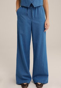 Široké modré džíny s páskem na stažení, nošené s krátkým topem a plochými botami s leopardím vzorem.