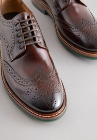 Brune lædersko med brogue-detaljer, forlænget snude og brune snørebånd. Har en kontrasterende grøn gummisål med synlige syninger.