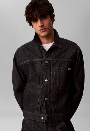 Junger Mann mit dunklem lockigem Haar, der eine schwarze Jeansjacke und passende Hose vor einem einfarbigen grauen Hintergrund trägt.