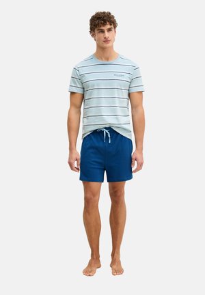 Jonge man die op blote voeten staat, gekleed in een lichtblauw gestreept t-shirt en marineblauwe short met trekkoord, tegen een eenvoudige achtergrond.