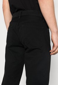 Jeans in cotone nero con un design classico a cinque tasche, passanti per la cintura e tessuto testurizzato, mostrati da dietro con dettagli di cucitura visibili.