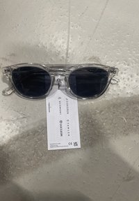 Gafas de sol de montura transparente con lentes oscuros, con forma de ojo de gato. Las varillas son delgadas y curvadas, descansando sobre una etiqueta con detalles de la marca.