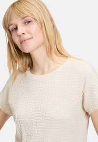 Jasno beżowy top o fakturowanej powierzchni, lekko prążkowanej, okrągłym dekolcie i krótkich rękawach. Cechuje się miękkim i luźnym krojem.