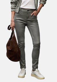 Persoon in grijze slim-fit broek, witte sneakers, een grijze jas, en met een bruine suède handtas aan de zijkant.