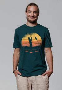 T-shirt en coton bleu canard foncé avec une impression graphique. Présente un coucher de soleil orange avec la silhouette d'un pêcheur et des illustrations de poissons en dessous.