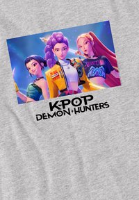 Grey t-shirt featuring a graphic of three animated characters with colorful hair, swords, and casual outfits; text reads "KPOP DEMON HUNTERS."
