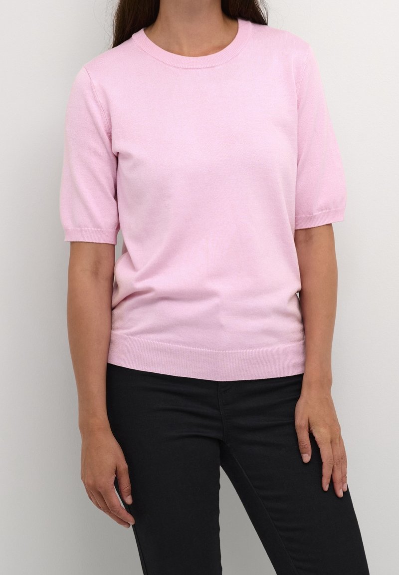 Femme portant un haut en maille rose clair uni à manches courtes et un pantalon noir, debout contre un mur blanc.