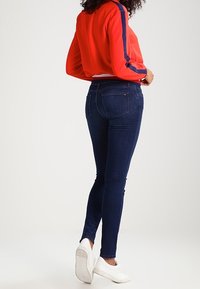 Figursydda mörkblå jeans med en slät textur, kombinerade med en röd topp med blå ränder och vita sneakers. Bakifrån sedd.