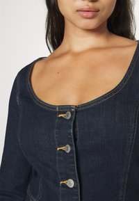 Gros plan d'une femme portant un haut en denim bleu foncé avec des boutons argentés et des coutures jaunes, montrant le décolleté et le haut de la poitrine.