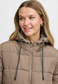 Veste rembourrée marron avec une fermeture éclair sur le devant, capuche à cordon élastique et cordon rayé noir et blanc contrastant. Texture lisse, design matelassé.