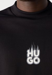 Gros plan sur une chemise noire avec le logo texte blanc "HUGO" conçu avec un effet de flamme sur la zone de la poitrine sous le col.