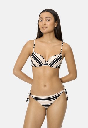 Mujer con bikini beige y negro a rayas, con braguitas de lazo a los lados, de pie con las manos detrás de la espalda contra un fondo claro y liso.