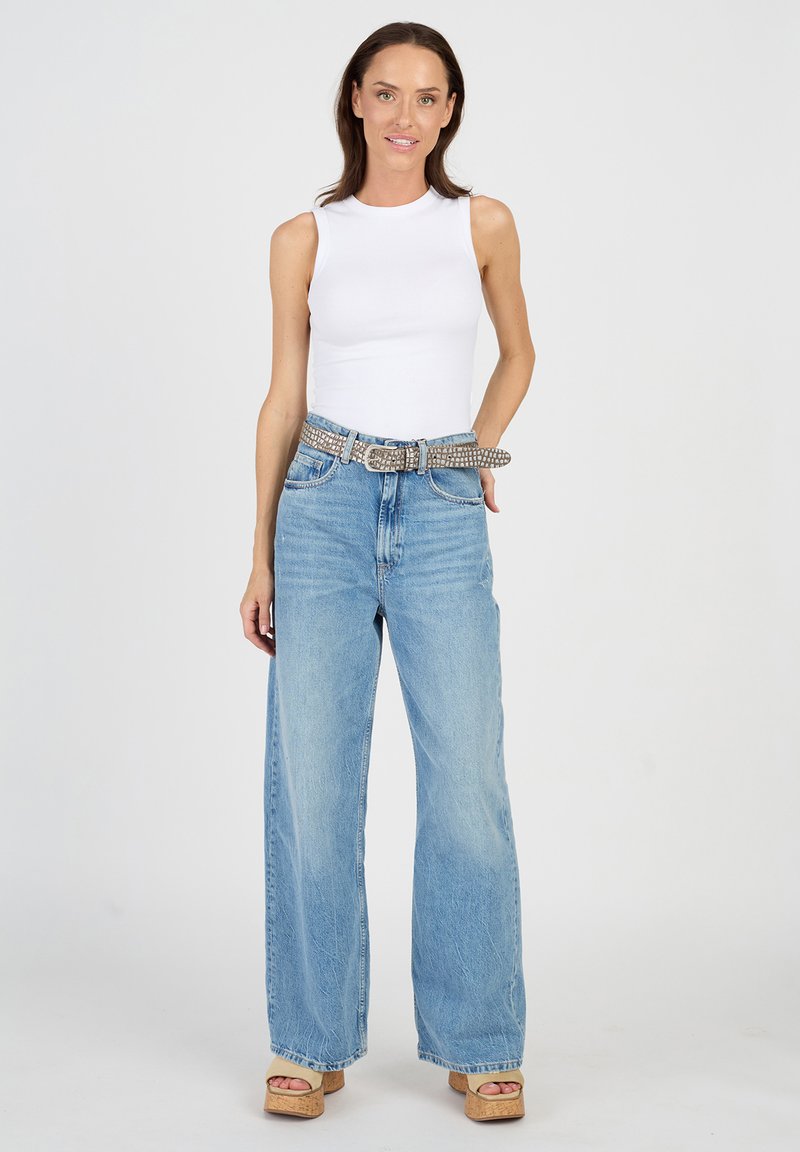 Valkoinen hihaton paita yhdistettynä vaaleansinisiin leveälahkeisiin farkkuihin ja kimaltavaan hopeanväriseen vyöhön. Malli seisoo korkean korkean sandaalin päällä.