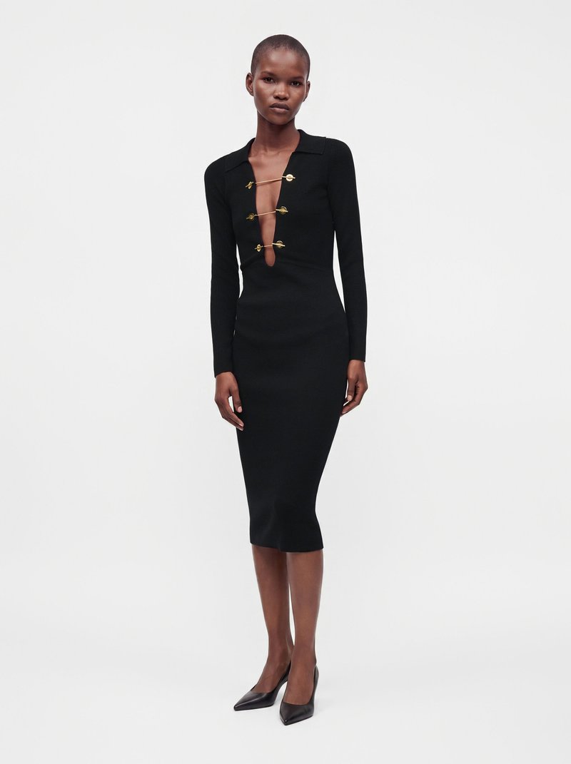 Femme portant une robe midi noire ajustée avec des manches longues et des fermoirs dorés le long d'une fente profonde à l'avant, associée à des talons pointus noirs.