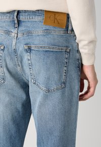 Jean en denim bleu clair avec une étiquette en cuir Calvin Klein à la ceinture, cinq poches et une texture délavée avec des coutures visibles.