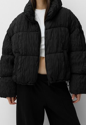 Femme portant une doudoune noire texturée par-dessus un haut court blanc et un pantalon noir, sur un fond gris uni.
