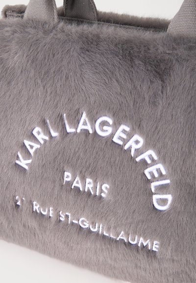 Крупен план на сива кожена чанта с черни тъкани дръжки и бял надпис Karl Lagerfeld Paris.
