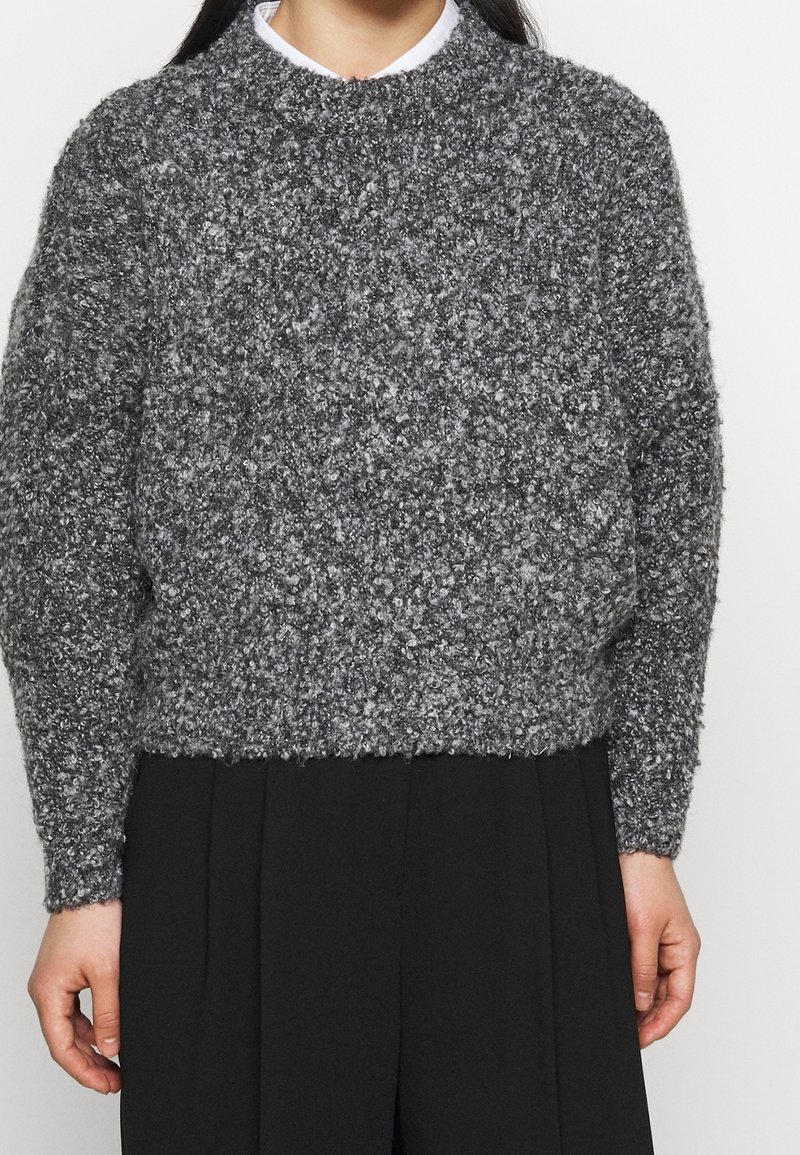 Personne portant un pull en maille gris texturé sur une chemise blanche à col et un pantalon noir ample, visible des épaules aux hanches.