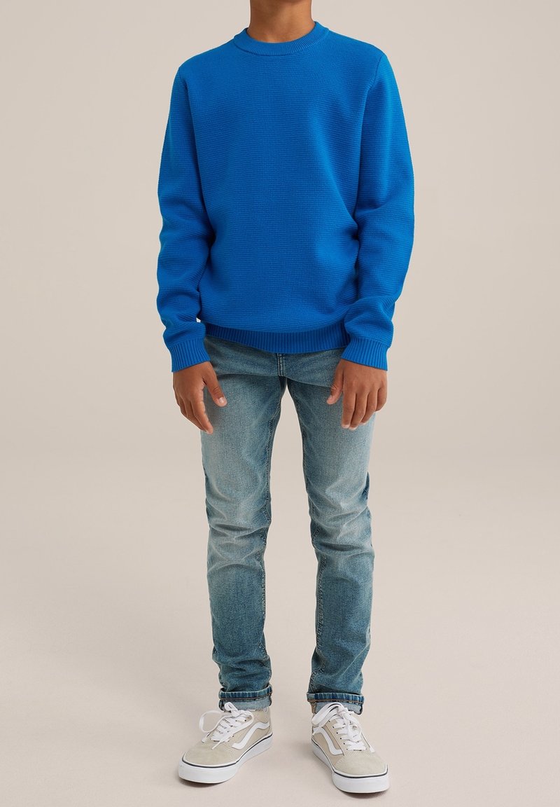 Enfant portant un pull bleu vif, un jean bleu clair et des baskets beiges, debout devant un fond clair.