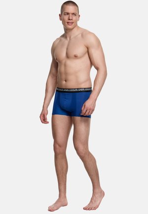 Sportlicher junger Mann, barfuß stehend, trägt blaue Boxershorts mit schwarzem Bund vor schlichtem weißem Hintergrund.