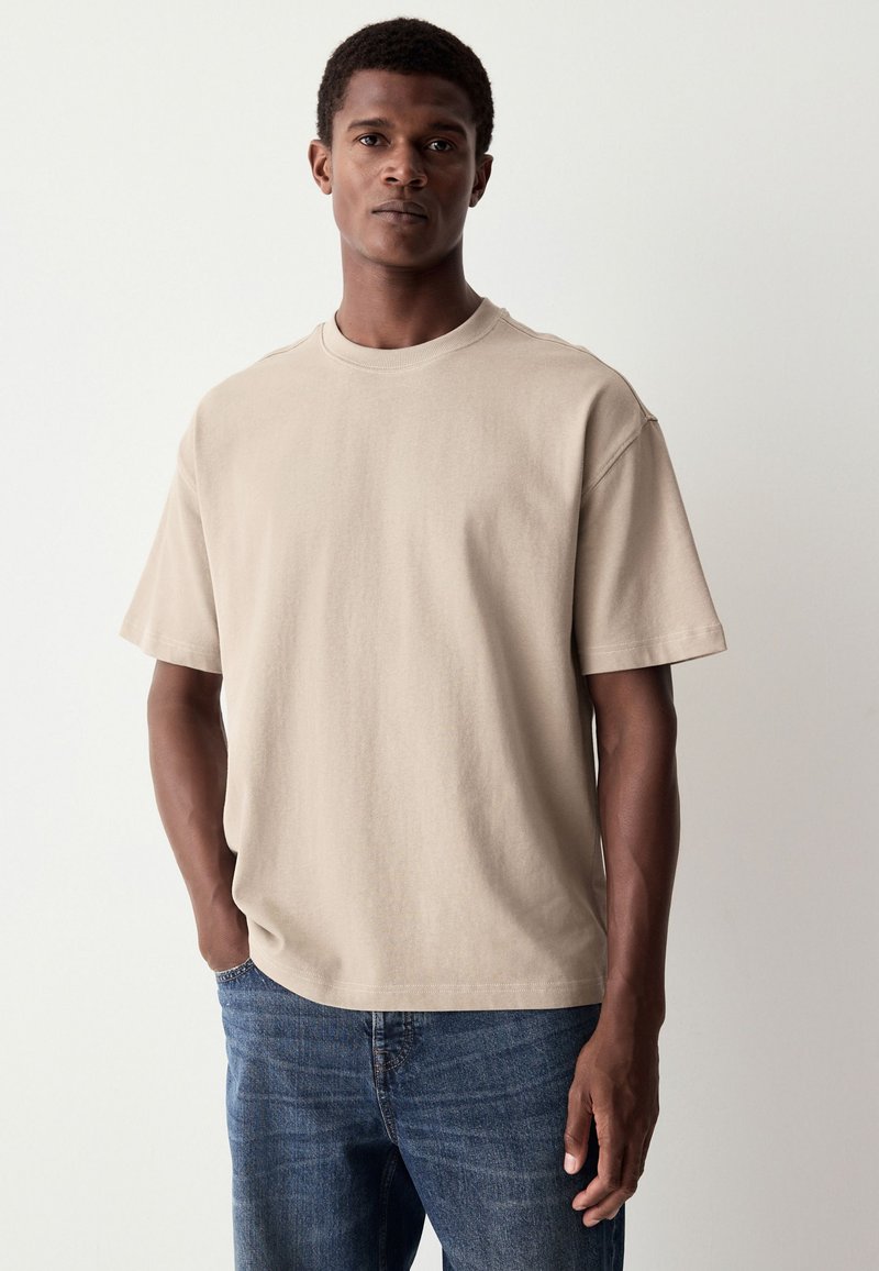 Beige katoenen T-shirt met korte mouwen en een ronde halslijn, gecombineerd met blauwe spijkerbroek. Eenvoudig ontwerp zonder zichtbare patronen of logo's.